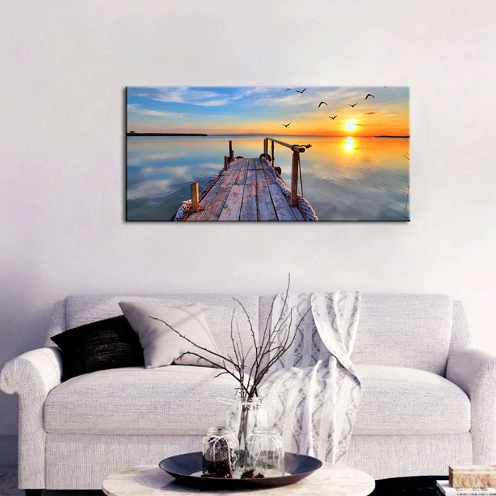 Wooden pier leading into calm sea at an orange sunset, canvas painting above a white sofa